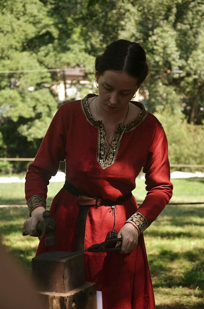 Woman in medieval dress hammers metal outdoors during reenactment.